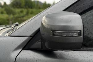 cerrar el material de la cubierta del ala del espejo lateral del automóvil gris es tiempo de estacionamiento plegable. por automático del sistema. en la superficie del coche, el agua mojada son gotas de lluvia del estacionamiento al aire libre mientras llueve. foto