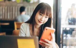 joven estudiante asiática adulta que usa un teléfono móvil para una aplicación en línea en un café interior.