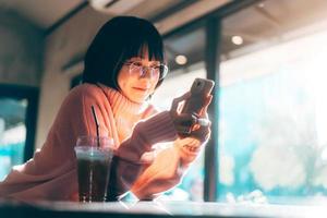 mujer asiática adulta joven que usa teléfono móvil para los medios sociales con ventana y luz solar cálida en invierno