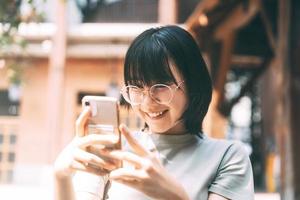 una joven adulta feliz asiática usa anteojos usando un teléfono móvil para las redes sociales.