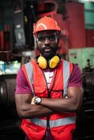 retrato de ojos sobrios y decididos trabajador afroamericano tiene barba descuidada en uniforme de seguridad con casco, gafas, chaleco y guante en la fábrica industrial. foto