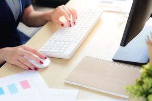 cerrar las manos de la mujer oficial con la mano derecha sostenía el mouse y la izquierda está escribiendo en el teclado blanco de la computadora con documentos de datos y cuaderno en el escritorio de la oficina moderna foto