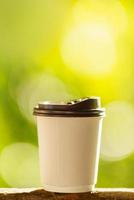 Closeup of disposable take away paper cup of hot coffee on wooden bar with green nature background.