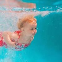Smiling baby girl in cute modern dress diving underwater in blue swimming pool. Active lifestyle, child swimming lesson with parents. Water sports activity during family summer vacation in resort photo