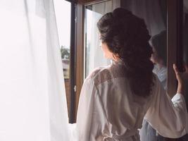 portrait of beautiful happy cheerful brunette woman in white silk robe standing by the window in the morning photo