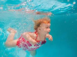 Smiling baby girl in cute modern dress diving underwater in blue swimming pool. Active lifestyle, child swimming lesson with parents. Water sports activity during family summer vacation in resort photo