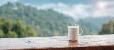 taza blanca de café o té caliente sobre una mesa de madera por la mañana con fondo de montaña y naturaleza foto