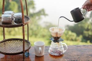 verter agua caliente a mano durante la preparación de café arábica con un equipo de goteo de café antiguo en una mesa de madera por la mañana con fondo de montaña y naturaleza foto