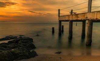 A long exposure landscape of beautiful sunset at the sea beach near concrete bridge with orange sky, clouds and stone