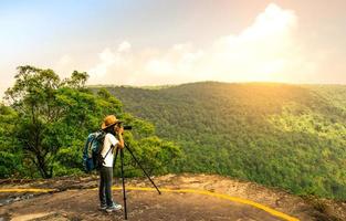 joven mujer viajera con sombrero de mochila y cámara en soporte de trípode en la cima del acantilado de la montaña viendo hermosas vistas de los bosques y el cielo en sus vacaciones. mujer asiática viaja sola. foto