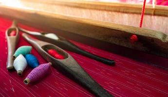 Selective focus on weaving shuttle tool on weaving machine. Red textile fabric weave. Weaving using traditional weaving loom and shuttle. Textile or cloth production in Thailand. Red handmade cloth. photo