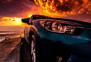 Blue compact SUV car with sport, modern, and luxury design parked on concrete road by the sea at sunset. Front view of beautiful hybrid car. Driving with confidence. Travel on vacation at the beach. photo