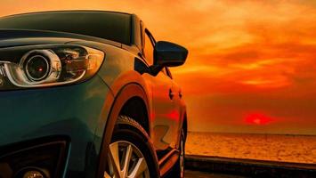 camión suv compacto azul con diseño deportivo y moderno estacionado en una carretera de hormigón junto a la playa al atardecer. vista frontal del coche de lujo. nuevo coche todoterreno con un hermoso cielo rojo al atardecer y nubes en la playa. foto