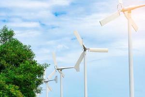 Horizontal axis wind turbine with blue sky and white clouds near green tree. Wind energy in eco wind farm. Green energy concept. Renewal energy. Alternative electricity source. Sustainable resources. photo