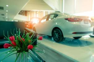 Fake red flowers in vase decorated on table in reception area in showroom. Artificial flower on blurred background of new and shiny white luxury car parked in modern showroom for sale. Car dealership. photo