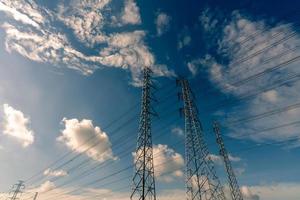 pilón eléctrico de alto voltaje y cable eléctrico contra el cielo azul y las nubes blancas. vista inferior del poste eléctrico con luz solar. torre de red de alto voltaje con cable de alambre. concepto de potencia y energía. foto