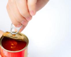 Woman's hand opens lid of canned fish on white background. Sardine in tomato sauce. Processed food. Canned fish in tin can. Seafood in can container. Preserve food. Tin can for storage preserve food. photo