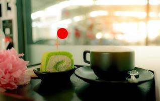 taza de café caliente con una rebanada de pastel de rollo suizo verde con crema en un plato en una cafetería en una bandeja de madera en la cafetería. café con leche, capuchino o espresso en vaso. bebida caliente. bebida cafeina para el desayuno foto