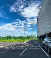 Car parking space at shopping mall for customer service. Outdoors asphalt car parking lot on sunny day with green tree forest and high voltage electric tower. Overhead transmission line. photo