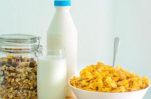 un vaso de leche y una botella de leche con una etiqueta en blanco colocada en una mesa de madera cerca de un tazón de cereal con una cuchara. desayuno de calcio para niños antes de ir a la escuela por la mañana. copos de maíz y concepto de leche foto