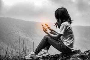 Black and white scene of young Asian woman sitting on the rock at the cliff and using smart phone. Adventure travel and communication concept photo