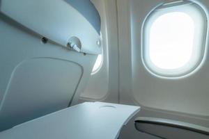 Plane window with white sunlight. Empty plastic airplane tray table at seat back. Economy class airplane window. Inside of commercial airline. Seat with armchair. Leather seat of economy class plane. photo