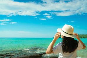 Happy young Asian woman in casual style fashion with straw hat stand at sea beach of resort in summer vacation . Relaxing and enjoying holiday at tropical paradise beach. Summer vibes. Summer travel. photo