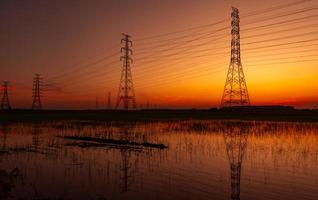 pilón eléctrico de alto voltaje y cable eléctrico con cielo al atardecer. postes de electricidad concepto de potencia y energía. torre de red de alto voltaje con cable de alambre en el campo de cultivo de arroz cerca de la fábrica industrial. foto