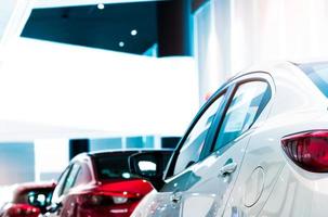 Rear view of white luxury car parked in modern showroom for sale. White shiny car on blurred red car in showroom. Car dealership. Coronavirus impact on automotive industry concept. Showroom interior. photo