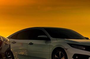 Front view of white car with luxury and modern design parked at parking lot with beautiful orange sunset sky. Automotive industry. Road trip travel. Parked car for watching sunset sky after work. photo