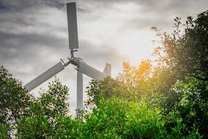 Horizontal axis wind turbine with blue sky and white clouds near green tree. Wind energy in eco wind farm. Green energy concept. Renewal energy. Alternative electricity source. Sustainable resources. photo