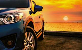 Blue luxury SUV car parked on concrete road by sea beach with beautiful red sunset sky. Summer vacation at tropical beach. Road trip. Front view sports and modern design SUV car. Summer travel by car. photo