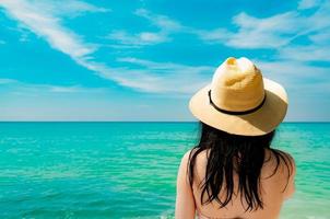 Vista trasera de una joven asiática sexy y feliz que usa un sombrero de paja para relajarse y disfrutar de unas vacaciones en la playa del paraíso tropical. chica de pie en la playa y mirando el mar en vacaciones de verano. vibras de verano. foto