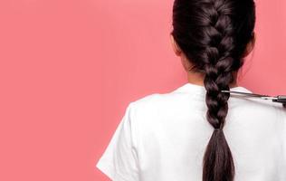 Back view of Asian woman with braided hairstyle is cutting with scissors for donate to cancer patients. Hair donation for breast cancer person. Woman with black long hair braid on pink background. photo