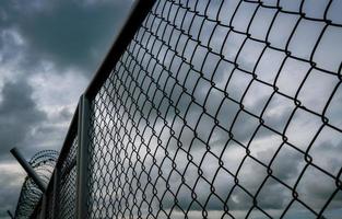 Military zone mesh fence. Prison security fence. Looking up view of barbed wire security fence with storm sky and dark clouds. Razor wire jail fence. Barrier border. Boundary security wall. photo