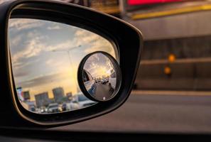 Reflection of traffic flow on asphalt road in side mirror of blue SUV. Car wing mirror with convex mirror for safety driving. View on the road and sunset sky behind cityscape through car side mirror. photo