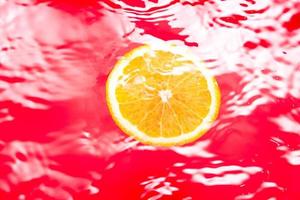 Piece of fresh orange on water surface with waves and reflections. Swimming piece of orange on red abstract background. photo