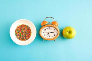 Alarm clock with a green apple and a bowl of sugar sprinkles on blue background. photo