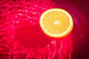 Piece of fresh orange on water surface with waves and reflections. Swimming piece of orange on red abstract background. photo