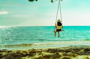 Asian woman in casual style wear hat swing the swings at sand beach  and looking beautiful tropical paradise sea and sky on sunset. Summer vacation. Summer vibes. Enjoying and relaxing girl on holiday photo