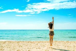 mujer joven feliz en camisas blancas y pantalones cortos saltando en la playa de arena. relajarse y disfrutar de unas vacaciones en la playa del paraíso tropical con cielo azul y nubes. chica en vacaciones de verano. vibras de verano. día feliz. foto