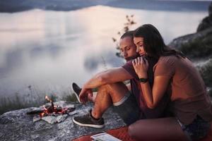 pareja abrazándose con mochila sentado cerca del fuego en la cima de la montaña disfrutando de la vista de la costa, un río o un lago. viajando a lo largo de las montañas y la costa, la libertad y el concepto de estilo de vida activo foto