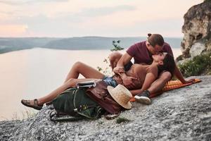 Hugging couple with backpack sitting on top of rock mountain enjoying view coast a river or lake. Traveling along mountains and coast, freedom and active lifestyle concept photo