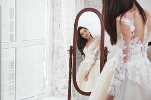 joven novia se mira en el espejo. el proceso de ajuste del vestido en el estudio de ropa artesanal foto