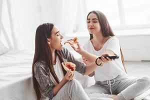 Taste mine slice. Sisters eating pizza when watching TV while sits on the floor of beautiful bedroom at daytime photo