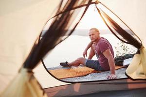 View from tent of traveller with backpack sitting on top of mountain enjoying view coast a river or lake. Traveling along mountains and coast, freedom and active lifestyle concept photo