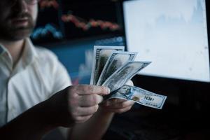 Counting the payment. Bearded man in white shirt counts money in the office with multiple computer screens in index charts photo