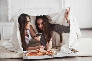 escondiéndose bajo una cálida manta. hermanas comiendo pizza cuando ven la televisión mientras están acostadas en el piso de un hermoso dormitorio durante el día foto