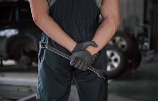 Mechanic looks forward. Rear view of worker in uniform that stands in workshop with wrench in hand photo