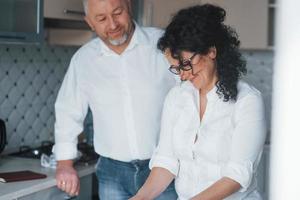 tener buen humor. hombre y su esposa con camisa blanca preparando comida en la cocina usando verduras